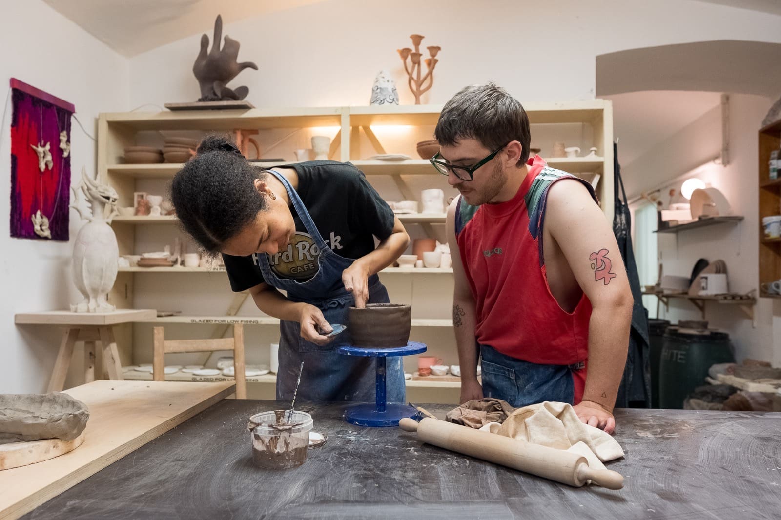 Two people at a work table, one smoothing a clay pot on a banding wheel